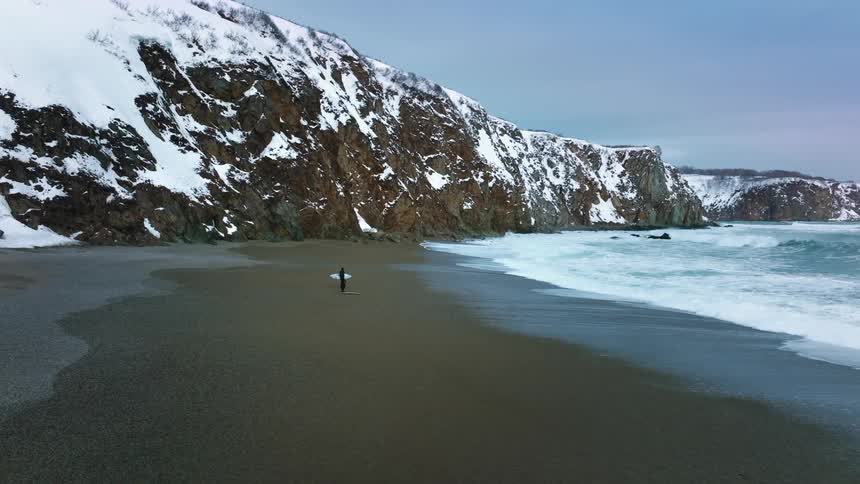 Two surfers wearing wetsuits stand on the beach near snow covered mountains watching waves roll into shore