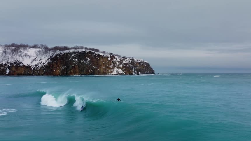 Two surfers riding large waves near a snow covered cliff taken from a distance