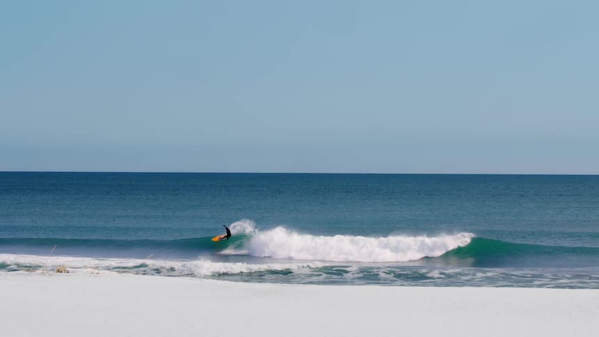 A surfer rides a perfect wave on a pristine beach with a clear blue sky overhead