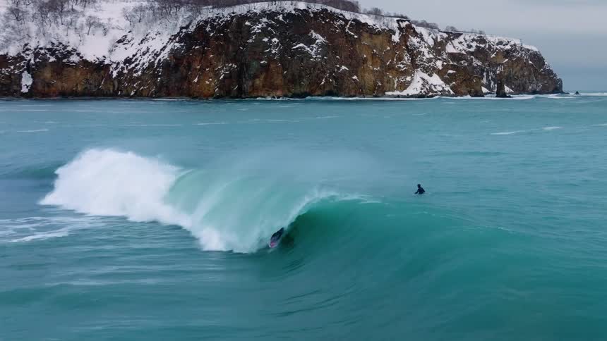A surfer in a wetsuit rides a wave with snowy cliffs in the background
