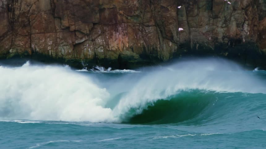 A powerful ocean wave crashes near a rocky cliff with mist spraying into the air and seabirds flying overhead