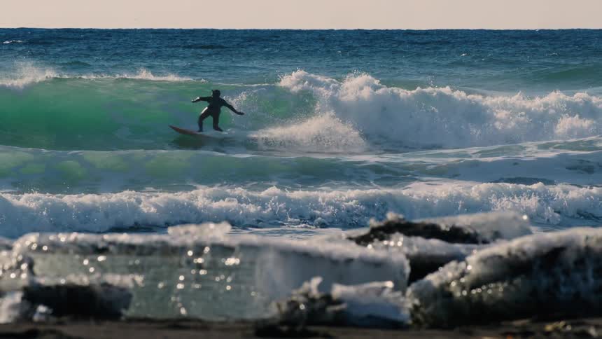 A surfer performing a powerful aerial maneuver over a large wave while in the foreground chunks of ice wash up on a rocky shore
