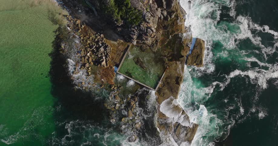 Aerial view of a natural pool on a rocky coastline with green ocean water and waves crashing