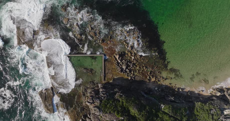 An aerial view of a natural pool on a rocky coastline surrounded by clear green water and tall waves