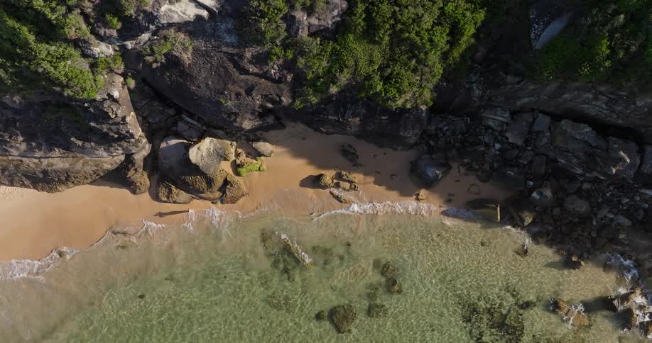 An aerial view of a rocky coastline with clear shallow water lapping against a small sandy beach