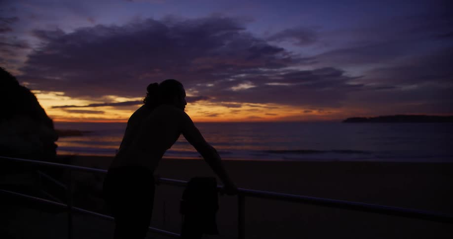 Man leaning on a railing facing a beach with the sunrise on the horizon