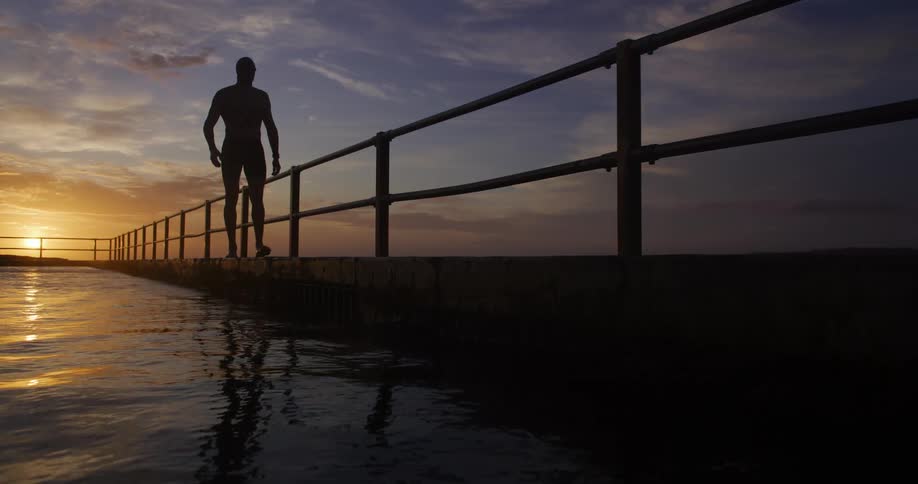 A silhouette of a person is walking along a pier with railings at sunset