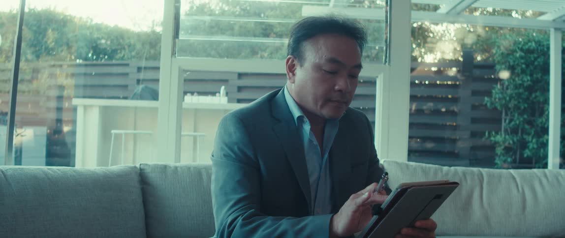 A man in a suit is sitting on a white sofa using his tablet and in his hand there is a pen