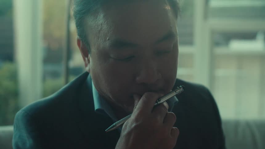 A middle aged asian man in a suit is thinking while holding a pen to his chin