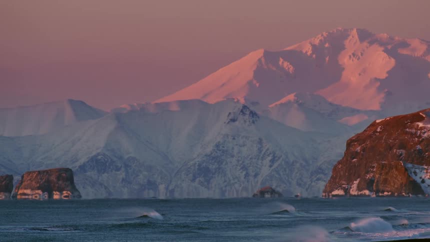 A snow capped mountain range bathed in the soft light of dawn reflected in the calm waters of the ocean