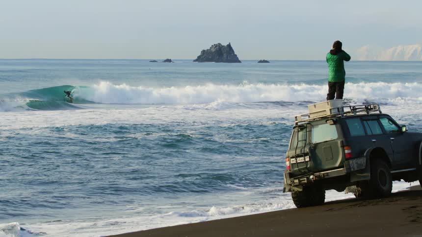 The film maker is filming a man surfing from the top of his vehicle