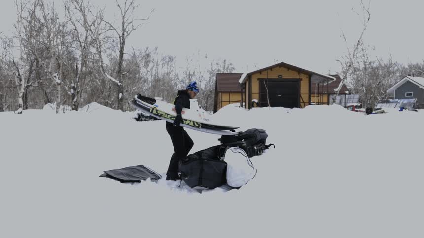 A man is packing a surfboard into black bags placed on the snow near a house in a snowy landscape