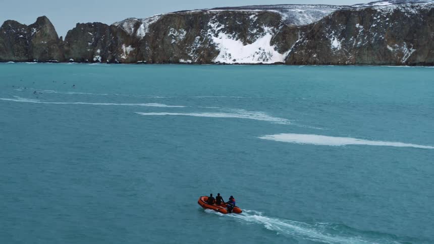 A small orange inflatable boat navigates through a vast icy blue sea with a snowcapped mountain range rising dramatically in the distance