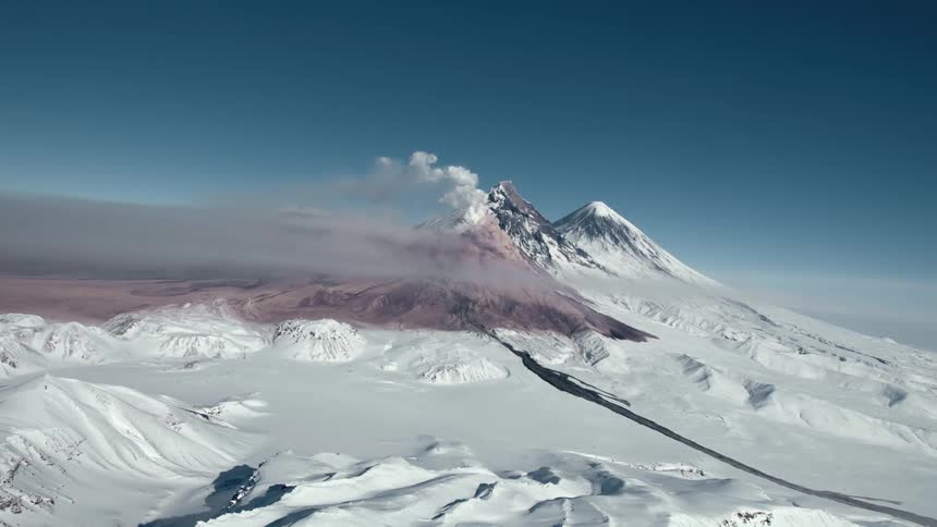 A majestic snowcapped volcano with a plume of smoke rising from its summit surrounded by a vast expanse of snowcovered mountains