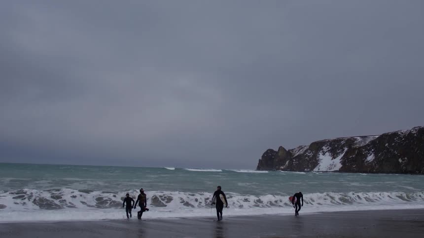Group of surfers coming out of the sea with rocky coast in the background with snow