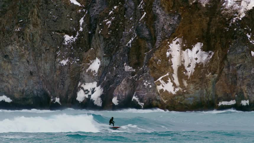 A man surfing over powerful waves crashing against a rugged snow covered cliff