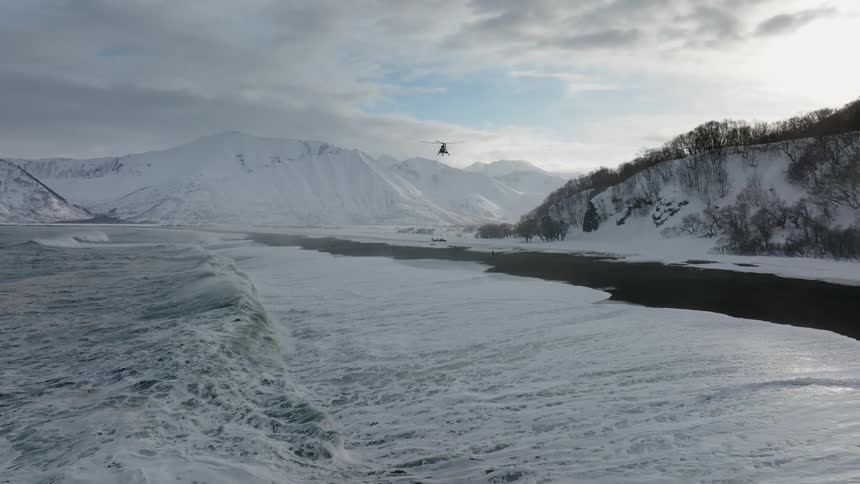 The shore of a beach with white waves and a helicopter flying overhead in the sky