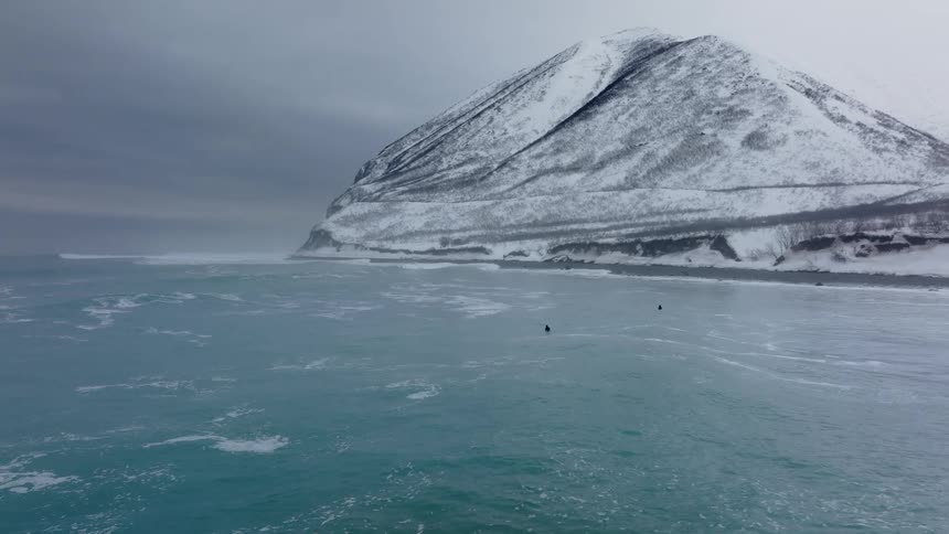 Two surfers ride waves in a cold icy ocean near a snow covered cliff