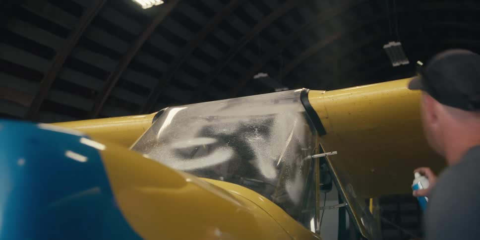 A man in a baseball cap sprays the windshield of a blue and yellow airplane inside a hangar