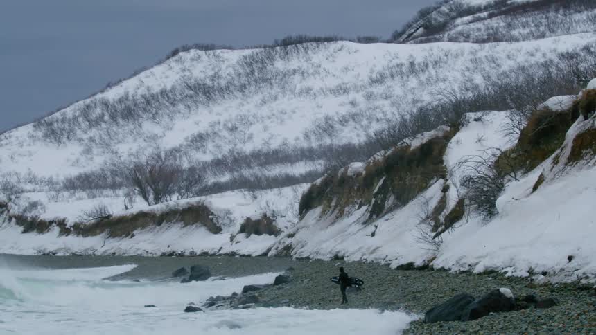A lone figure walking along a rocky beach in a snowy landscape with crashing waves and a steep snowcovered hillside in the background