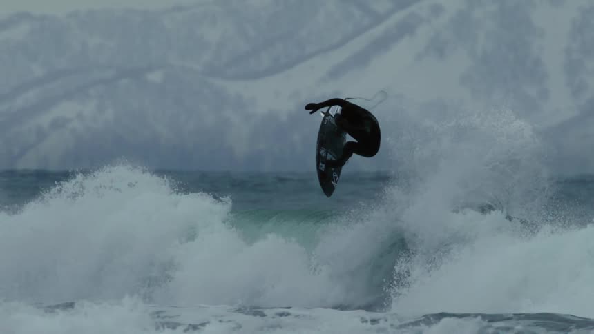 A person in a wetsuit is surfing on a wave in a cold snowy environment with mountains in the background