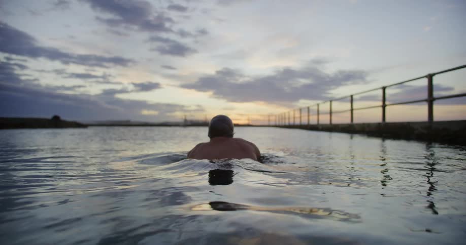 A person wearing a swim cap is swimming in an outdoors pool at sunset