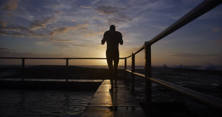 Man walking on a dock as the sun glows on the horizon and waves splash against the coast