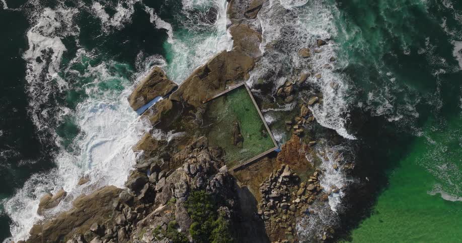 An aerial view of a natural rock pool surrounded by rugged rocks and crashing ocean waves