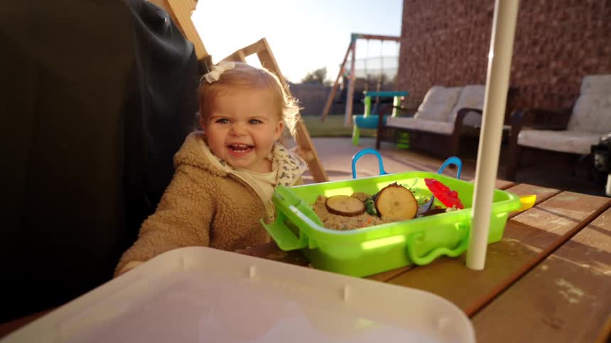 A toddler in a tan jacket smiles and laughs while playing at a table with a green container filled with sand and wooden pieces and a white tray of water nearby all outdoors