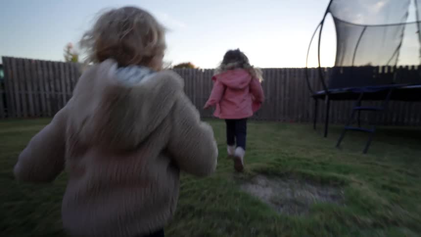 A little girl in a pink jacket runs around the backyard through a play area in the morning