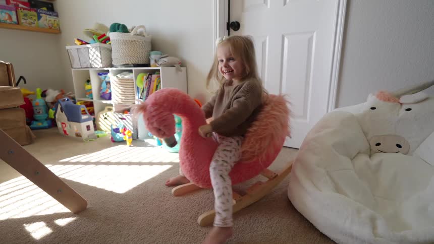 A child is playing on a pink flamingo rocking toy in a playroom filled with various toys and storage baskets