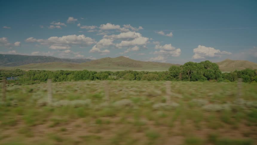 A dirt road cuts through a vast grassy plain framed by a distant mountain range under a bright blue sky