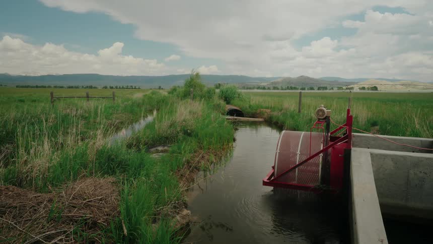 A small irrigation system with a red wheel and concrete structure sits at the edge of a narrow waterway flowing through a grassy field towards distant hills