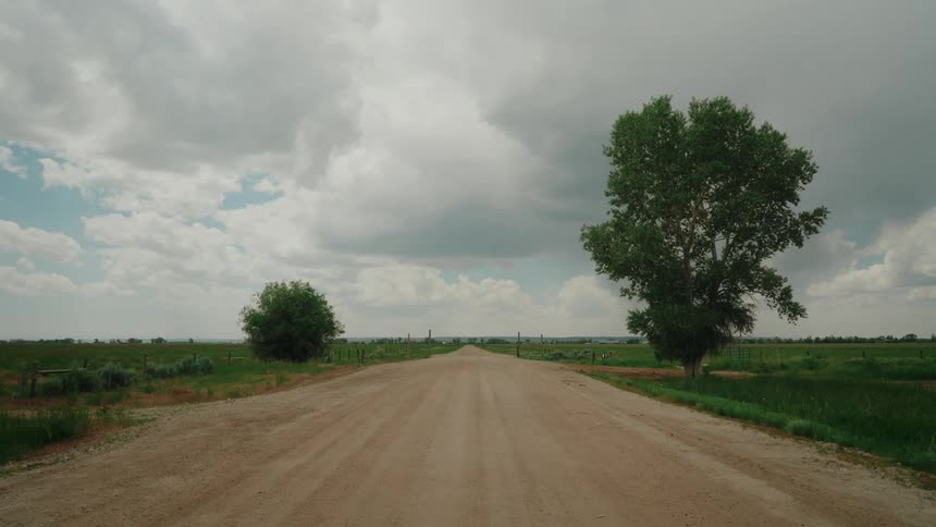 A long straight dirt road stretching into the distance under a dramatic sky with dark clouds passing through farmlands on either side