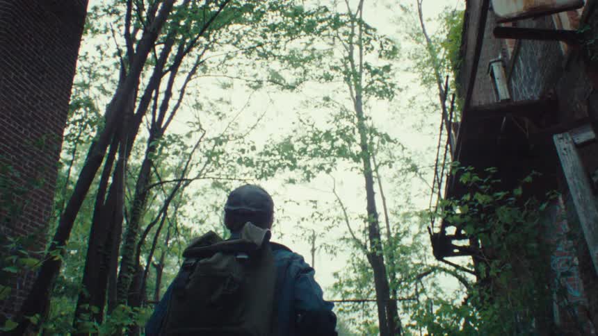 A person with a backpack and cap is walking through an overgrown abandoned area with graffiticovered brick walls and rusted metal structures