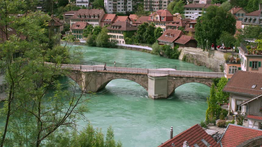 A picturesque city with colorful buildings lines a river with a vibrant green hue spanned by a historic stone bridge