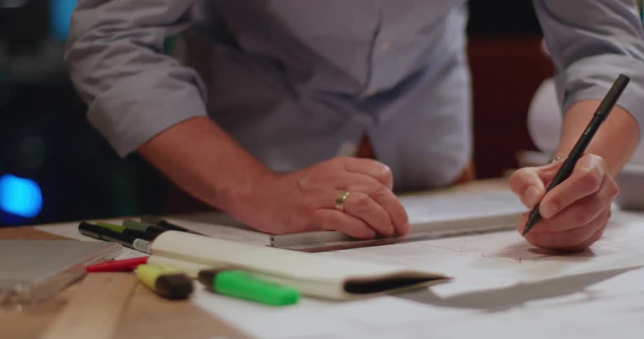 A person is using a ruler and pen to draw precise lines on a piece of paper with highlighters and other drawing tools nearby on the table