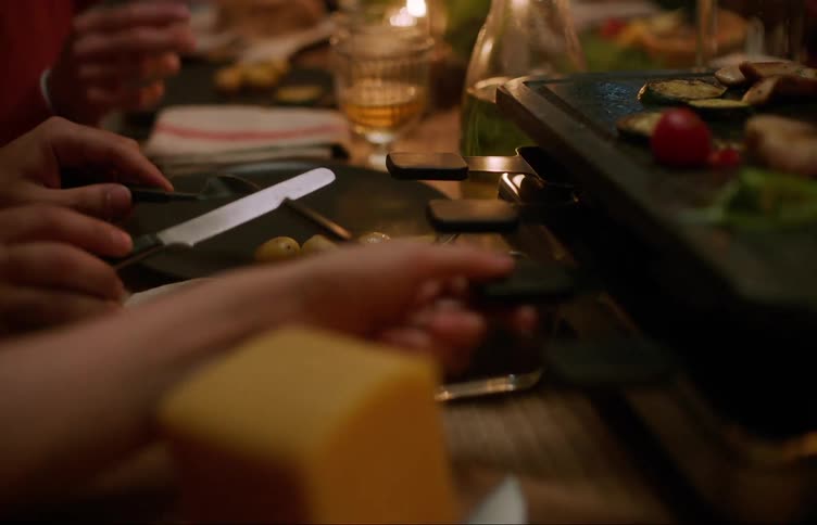 A person is preparing food on a raclette grill with a block of cheese and a knife