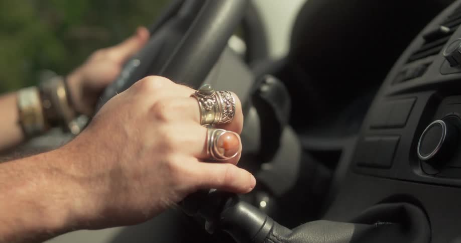 A hand adorned with ornate rings grips the steering wheel and gear shift while driving