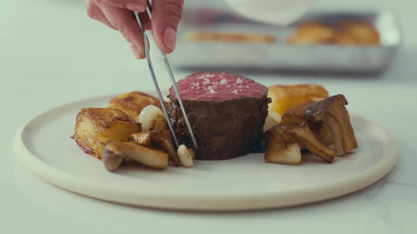 A hand using tongs arranges cooked mushrooms and potatoes around a seared steak on a plate