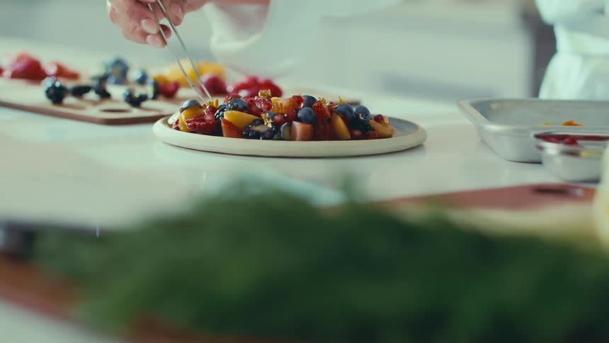 A person uses tweezers to arrange pieces of fruit on a plate with additional fruit on a cutting board in the background