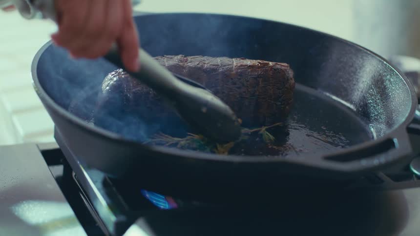 A large piece of meat being seared in a hot castiron pan on a stovetop with steam rising from the pan