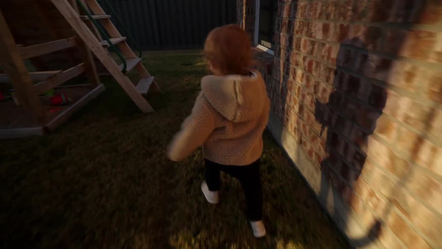 A little girl in a pink jacket runs around the backyard through a play area in the morning