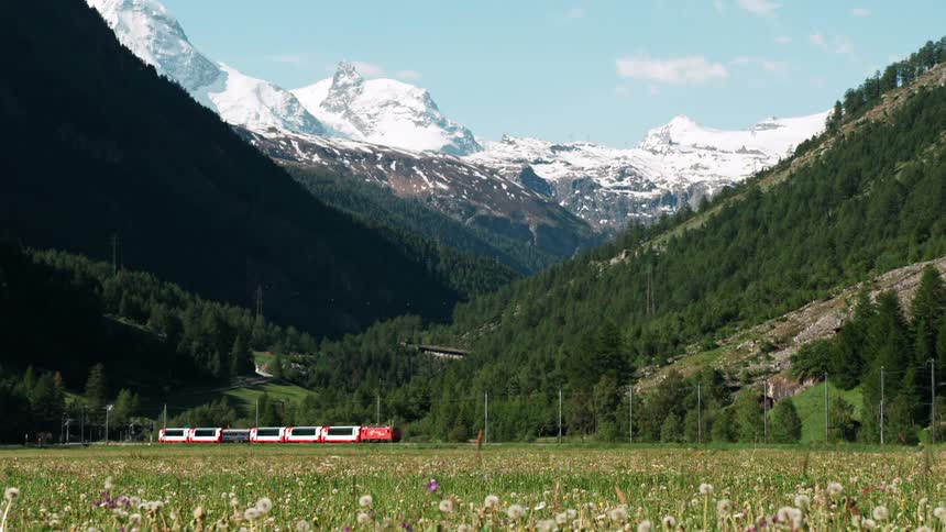 A red train traveling through a scenic valley surrounded by snowcapped mountains and lush green forests