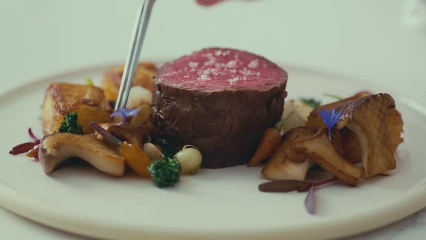 Hand with tweezers placing a purple flower on an elegantly assembled gourmet plate highlighting a juicy rare piece of meat sprinkled with coarse salt on top