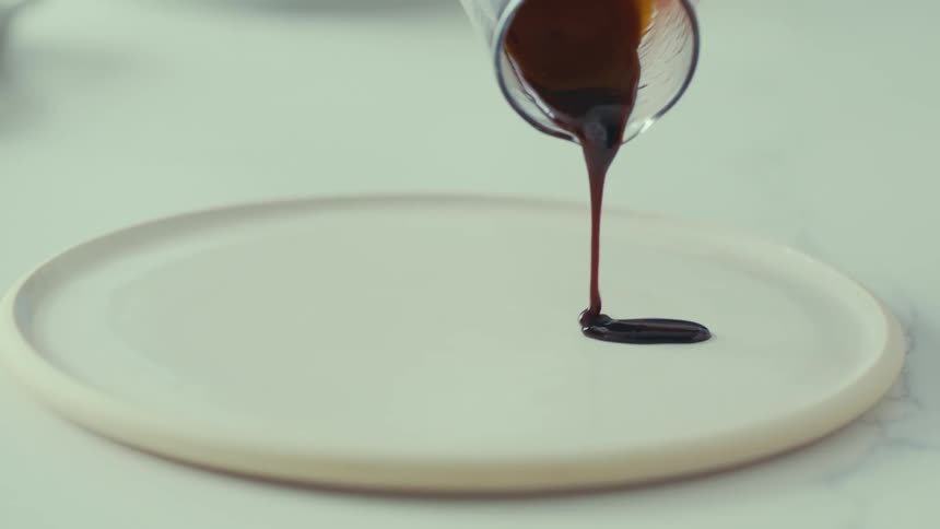 Dark liquid is being poured from a glass measuring cup onto a white plate