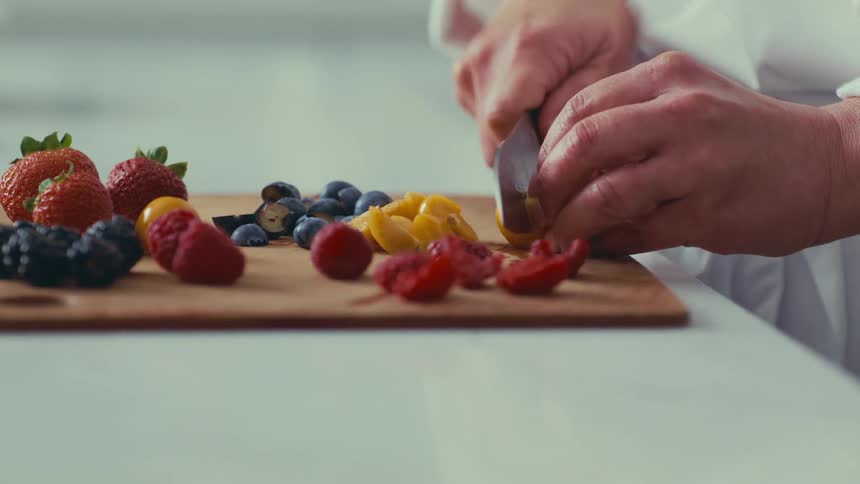 A person carefully slicing yellow cherry tomatoes on a wooden cutting board filled with a colorful assortment of fresh berries