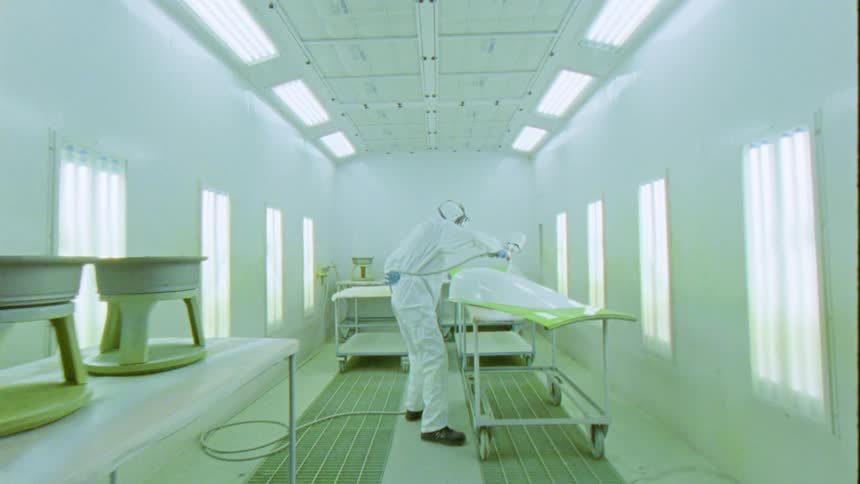 A person in white clothes and a protective mask using a machine to spray paint on a white metal plate