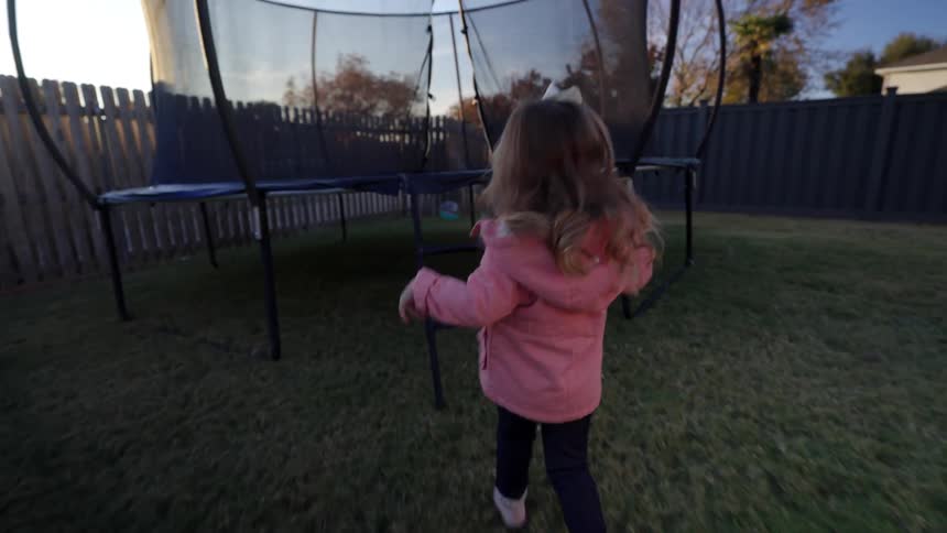 A girl wearing a pink jacket runs around a backyard and climbs into a trampoline in the morning