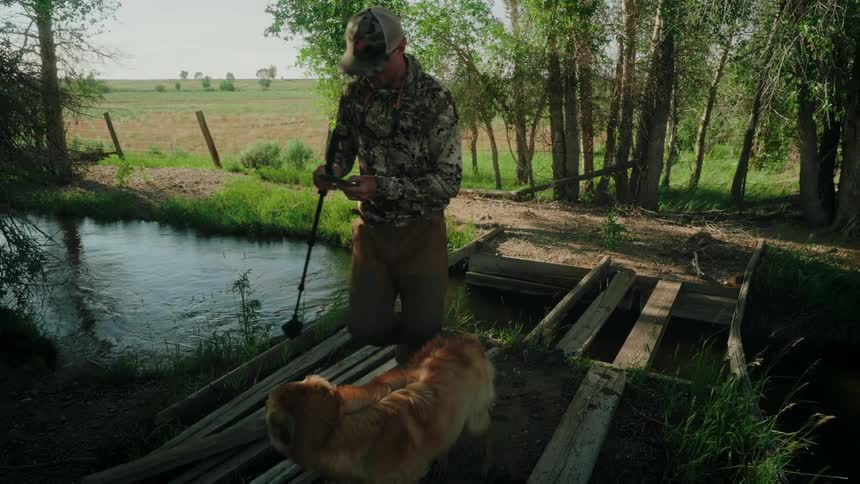 A man inspecting a decrepit wooden bridge over a stream in the countryside accompanied by a dog